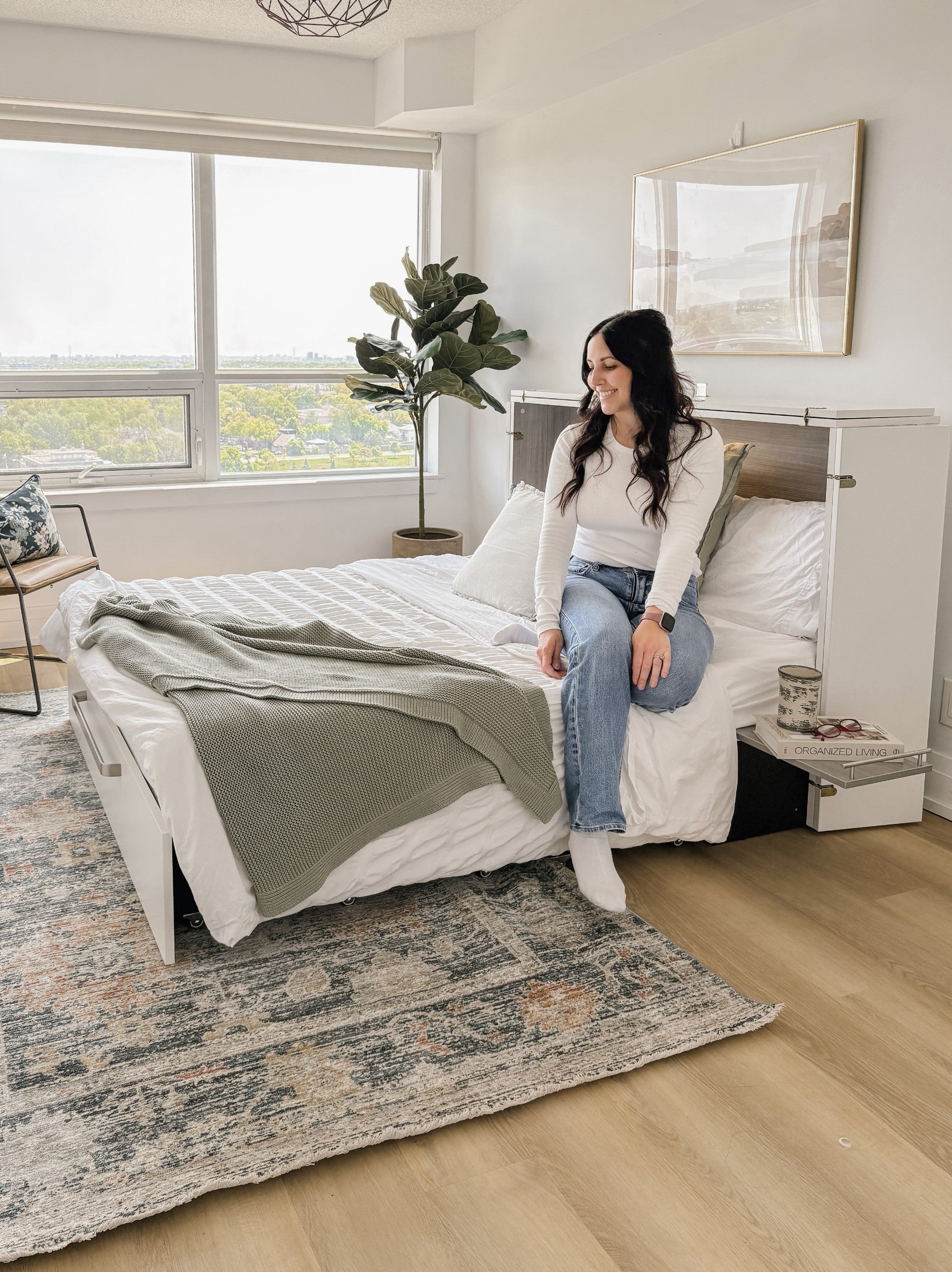 Woman sitting on a cabinet bed in a well-lit bedroom with large windows.