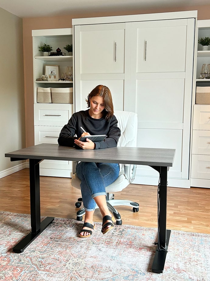 Person sitting at a desk using a tablet in a home office setting with white cabinets.