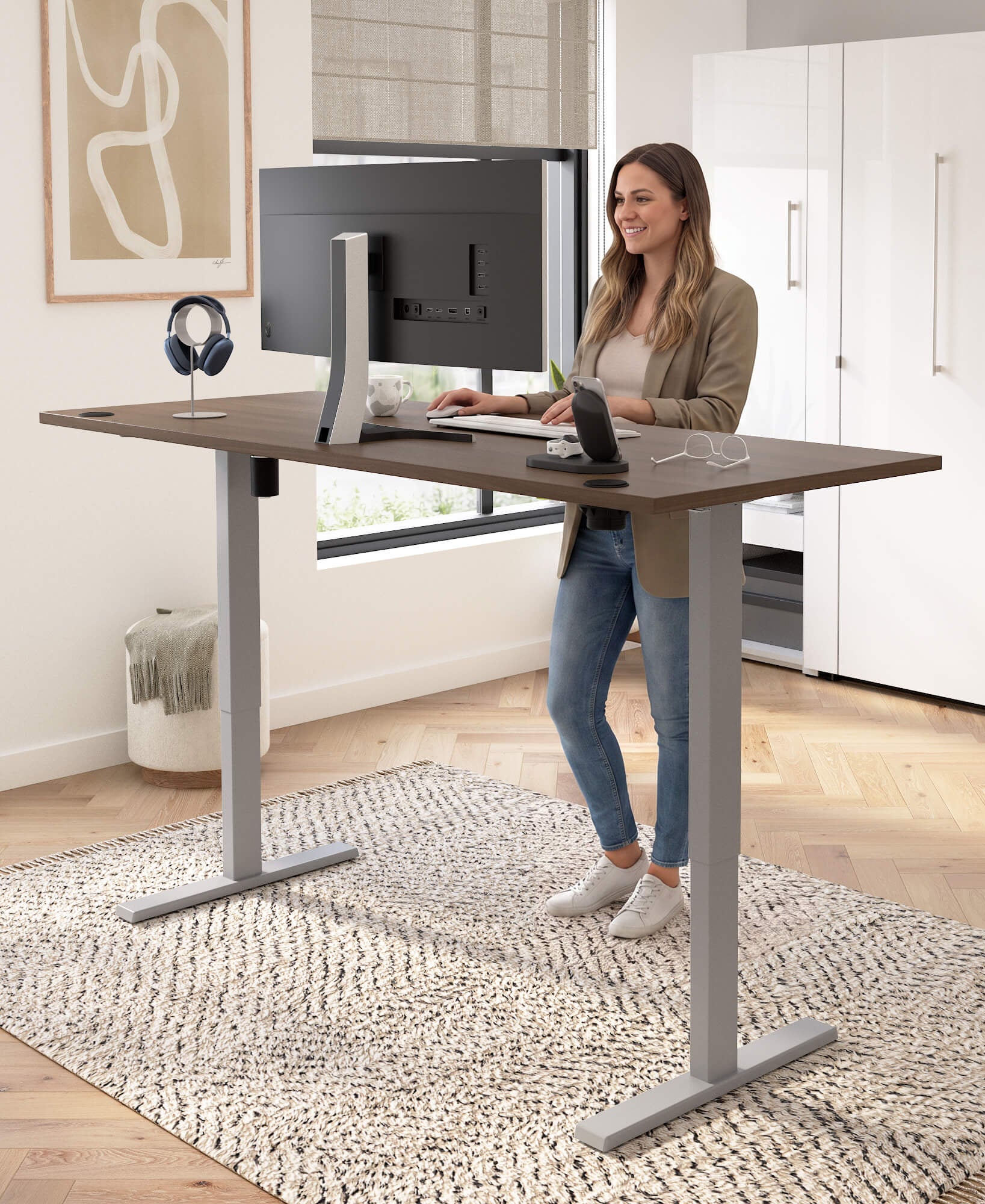 A woman stands at a height-adjustable standing desk in a bright home office. The desk has a dark brown wood-grain top and silver metal legs, with a computer monitor, keyboard, and headphones on top.