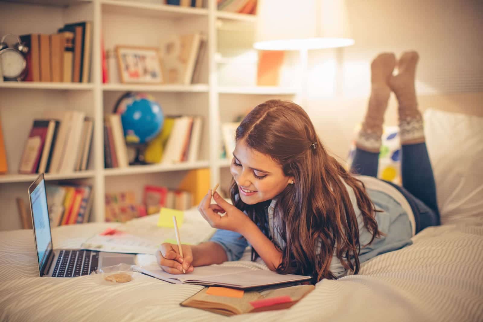 teenage girl laying on her bed studying with laptop and books