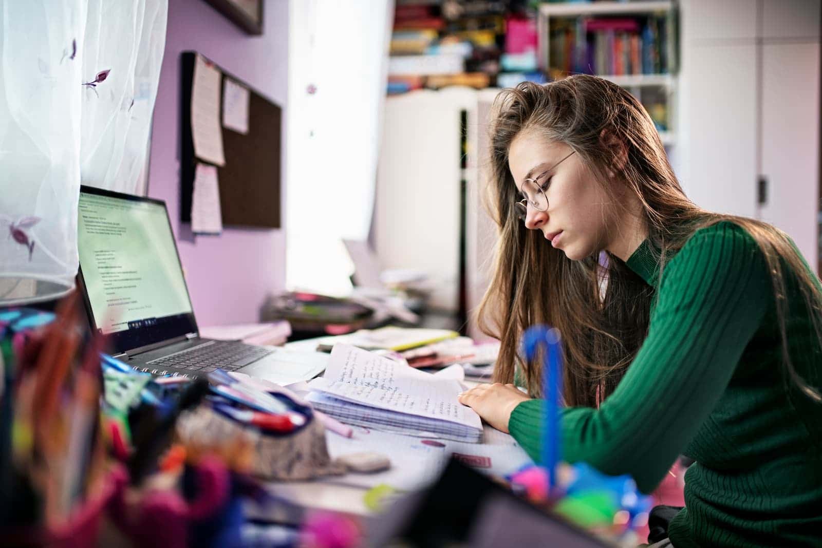 teenage girl sitting at desk with documents, laptop, and books. glasses on and post-its